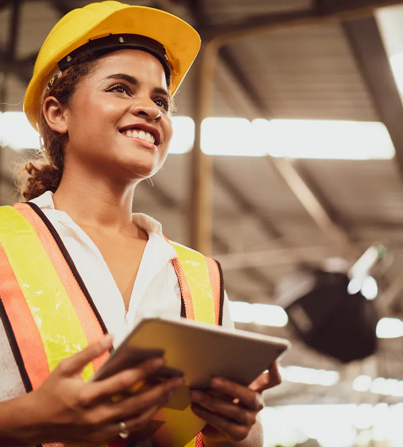 Smiling woman in yellow hard hat and safety vest holding a tablet in an industrial or construction setting.