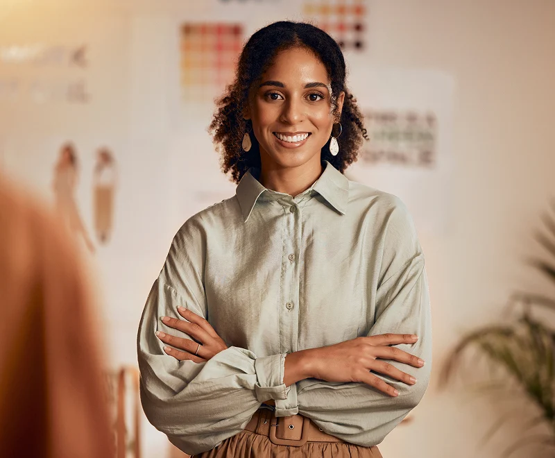 Professional woman with curly hair wearing a sage green button-up shirt, arms crossed, smiling at camera in an office or conference room setting.