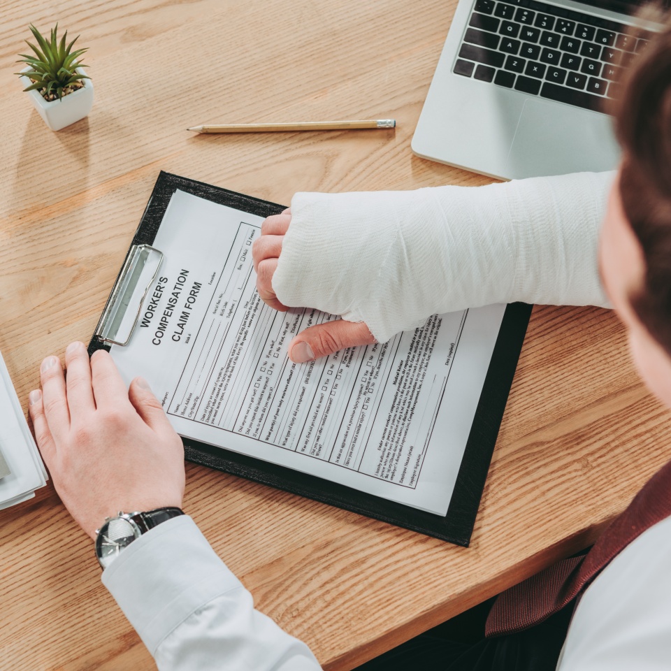 cropped view of worker holding broken arm compensation claim and sitting at table in office, compensation concept