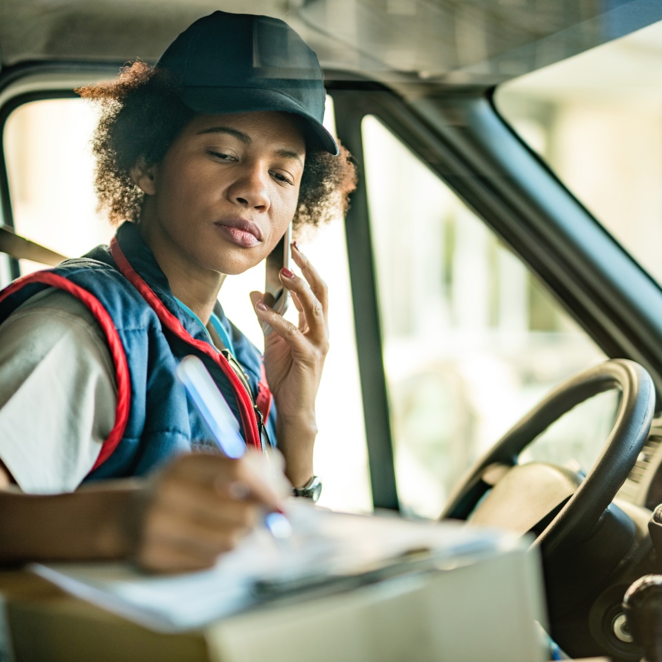 Female African American courier taking notes while talking on the phone in a delivery van.