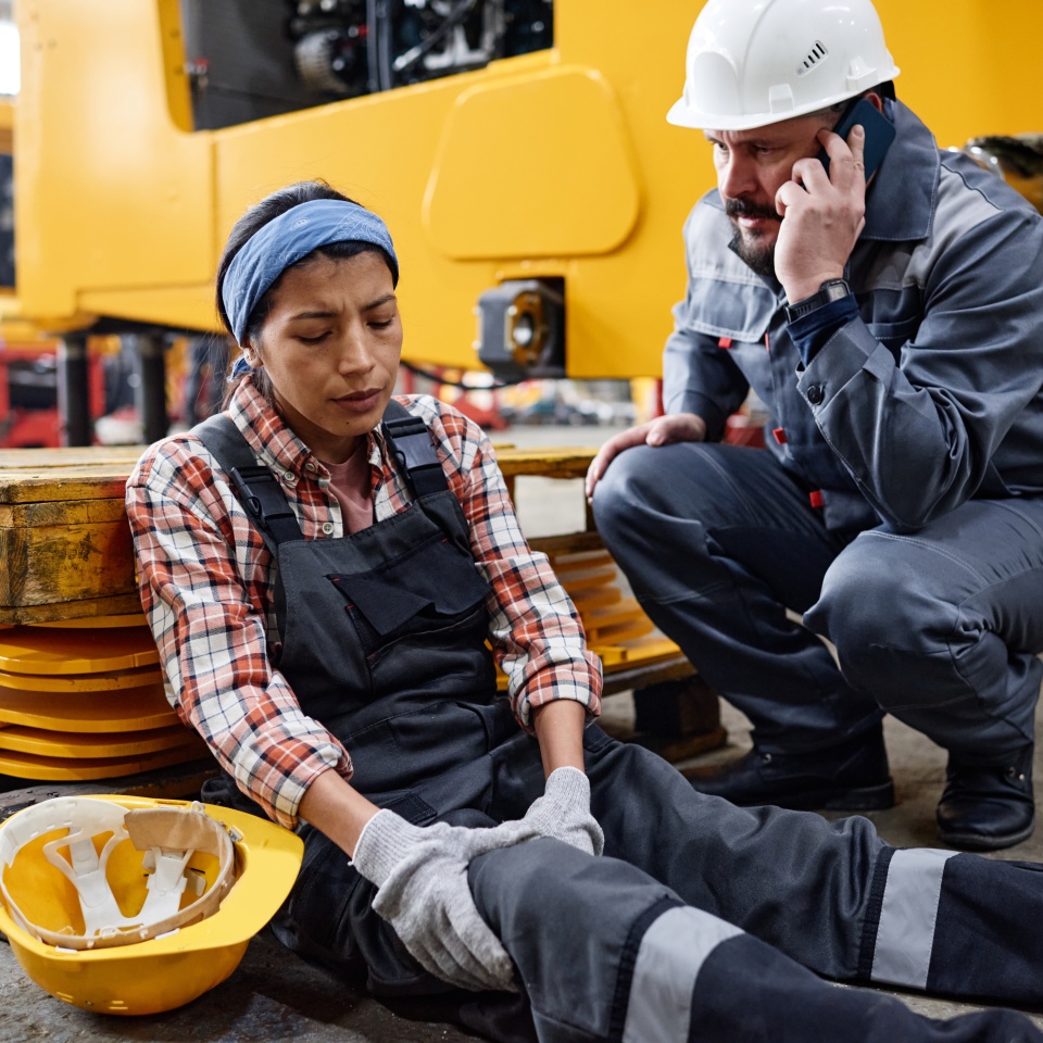 Young female engineer with hurting knee sitting on the floor by anxious male worker in safety helmet and uniform calling ambulance