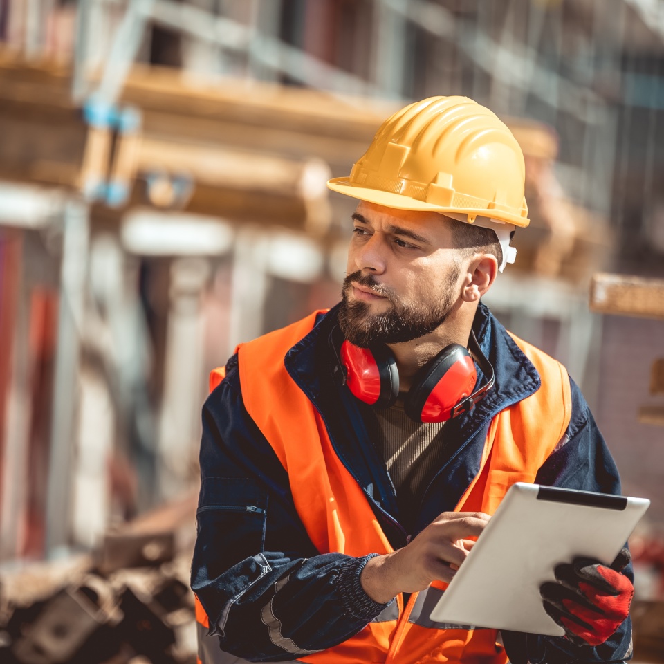 Construction site manager standing wearing safety vest and helmet,using his tablet at construction site.