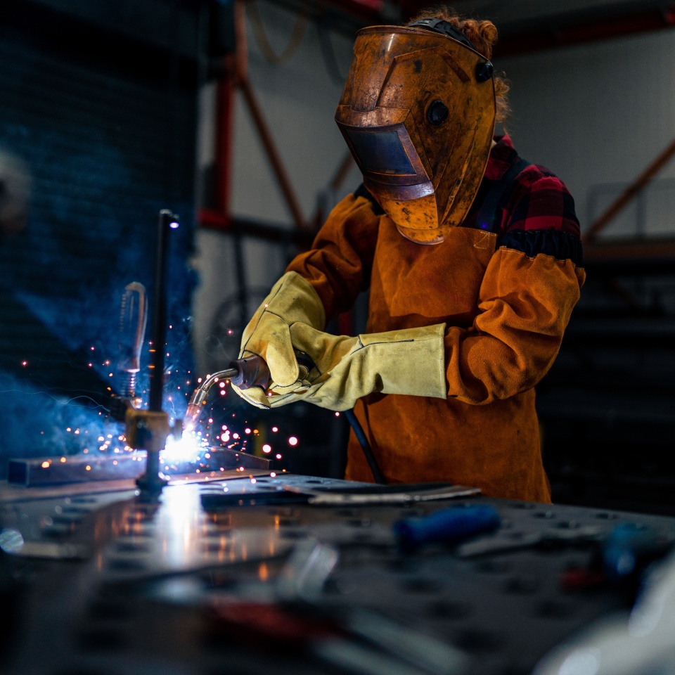 A woman in protective equipment uses tools and machines in the workshop, sparks fly and illuminate the workshop