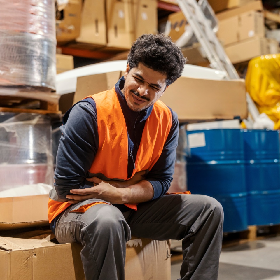 An interracial warehouse worker in pain sitting in storehouse surrounded by cardboard boxes and suffering from stomachache.
