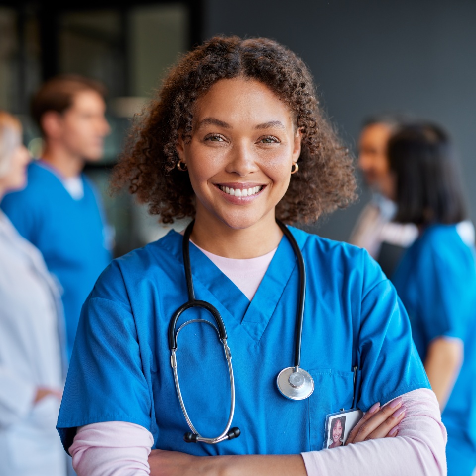 Confident happy woman nurse smiling with crossed arms with healthcare team in background. Multiethnic medical professional in scrub ready to assist patients. Portrait of reliable confident nurse.